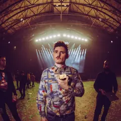 Dawid Podsiadło w futurystycznym, lekko cyberpunkowym wydaniu, z jabłkiem w dłoni, na tle hali koncertowej.