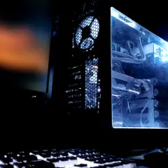 Komputer z podświetlanym wentylatorem i widocznymi podzespołami przez szklany panel. Na pierwszym planie klawiatura i myszka. W tle ekran z pomarańczowym gradientem. System UEFI gotowy do pracy.