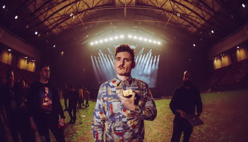 Dawid Podsiadło w futurystycznym, lekko cyberpunkowym wydaniu, z jabłkiem w dłoni, na tle hali koncertowej.