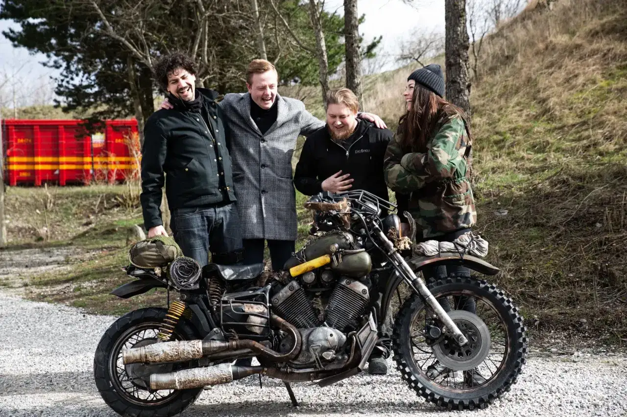 Czterech przyjaciół, jak z gry Days Gone 2, pozuje przy zmodyfikowanym motocyklu w postapokaliptycznym otoczeniu.